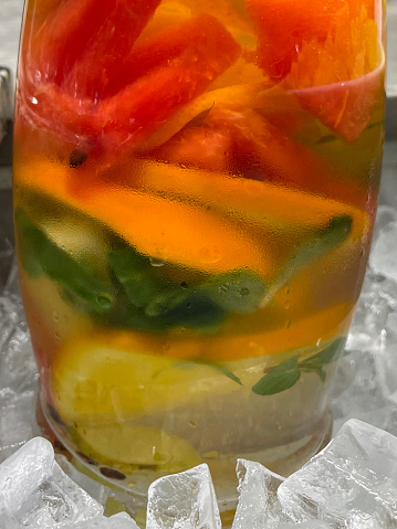Stock photo showing close-up view of layers of sliced fruit and vegetables in glass jug of detox drinking water surrounded by ice.