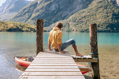 Man with red inflatable canoe on peaceful environment in Norway. Blue lake and green mountains.
He relaxes on a wooden pier.