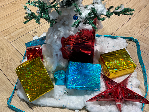 Stock photo showing close-up, elevated view of indoor, artificial Christmas tree covered in coloured baubles, with fake snow made of cotton wool and wrapped presents underneath on wooden flooring.