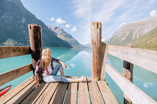 Woman with red inflatable canoe on peaceful environment in Norway. Blue lake and green mountains.\nShe relaxes on a wooden pier.