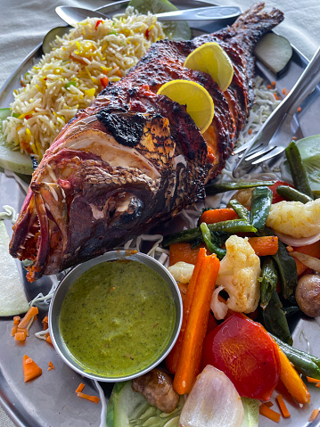 Stock photo showing close-up, elevated view of metal platter of tandoori marinated, grilled red snapper fish garnished with lemon slices and served on a bed of rice with vegetables.