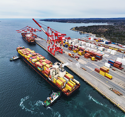 Aerial drone view of a container ship docking with the assistance of tugboats.