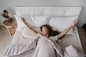 Young long haired Italian girl laying on bed eyes closed awakening at Sunday morning smiles satisfied by healthy sleep. Top view on young woman spreading hands, stretching. Cozy home, happy people.