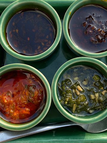 Stock photo showing a green square dish with four dipping sauces for dumplings in circular ramekins with a spoon for consumption, chilli oil, soya sauce, tomato and garlic spicy condiments, elevated overhead view