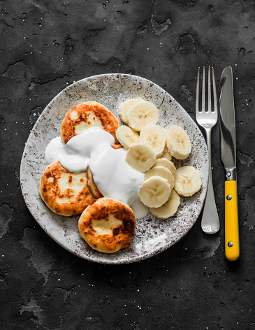 Cottage cheese syrnyky with greek yogurt and banana on a dark background, top view