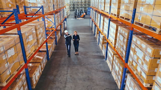 Young Caucasian female warehouse worker, mature adult woman manager working in factory warehouse, walk and discuss talk together. Logistic industry business, industrial job, or people at work concept