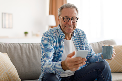 Portrait of smiling mature man in eyeglasses using mobile phone, watching video and holding cup of coffee, sitting on the couch in living room. Guy browsing internet, surfing web, free copy space