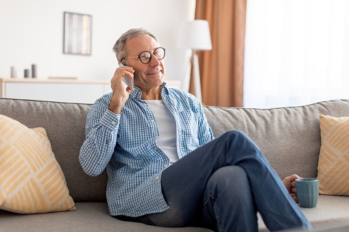 Portrait Of Happy Mature Man In Glasses Talking On Mobile Phone Sitting On Couch At Home, Drinking Coffee. Cheerful Adult Male Speaking, Having Cellphone Conversation. Modern Communication Concept