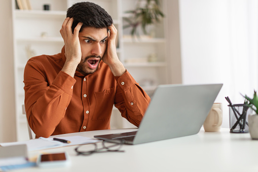 Oh No, Error Concept. Portrait of shocked scared Arab guy sitting at desk with laptop, screaming and grabbing his head. Remote Work. Stressed man suffering business problems, reading bad negative news