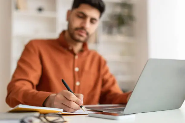 Business Education Concept. Focused young man sitting at desk working on laptop writing letter in paper notebook, selective focus on hand holding pen. Busy millennial male using pc, blurred background
