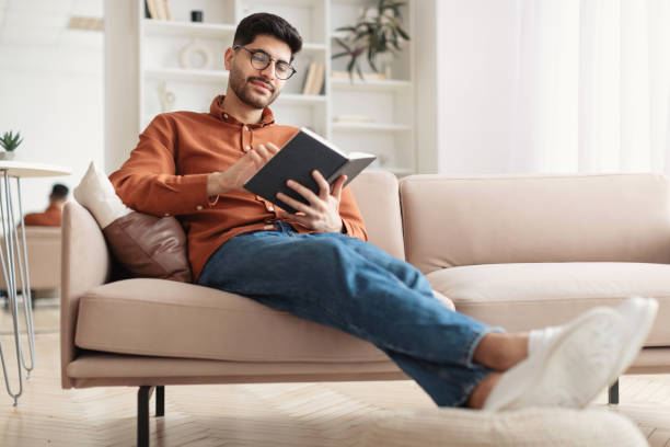 uomo arabo sorridente in occhiali che legge il libro di carta - reading book men sofa foto e immagini stock