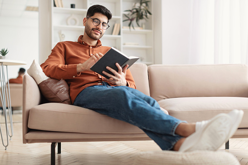 Leisure and Relax. Smiling young Middle Eastern man wearing eyeglasses holding paper book, sitting on couch at home in living room, reading literature, enjoying story, full body length portrait