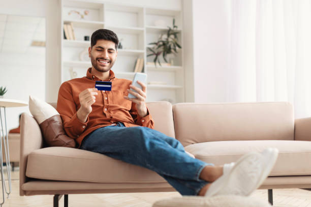 istock Smiling Arab guy using phone and credit card at home 1477233969