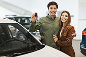 Joyful young Caucasian couple holding new car key, smiling at camera in modern dealership store