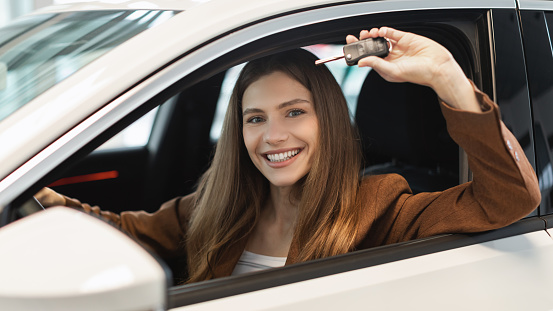 Happy young woman showing car key from auto window, purchasing new vehicle at showroom store, banner. Cheery millennial female client sitting in drivers seat, buying luxury automobile at dealership