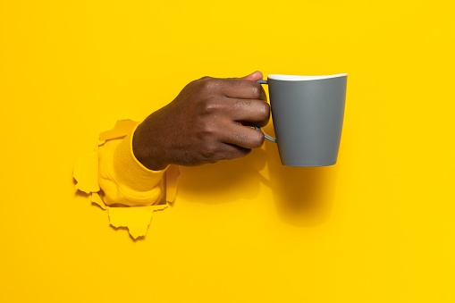 Black male hand holding a mug of coffee through torn yellow paper background, closeup, copy space. Have a break and refreshing drink