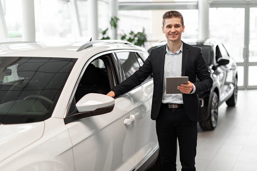 Cheerful young sales manager with tablet computer standing near new automobile at luxury car salon, copy space. Millennial insurance agent at work in modern automotive dealership store