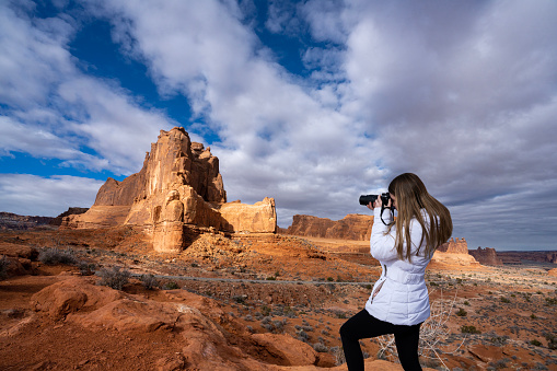 Woman taking photo on hiking trail, snowy mountains in the background.  Arches National Park. Moab Utah, USA