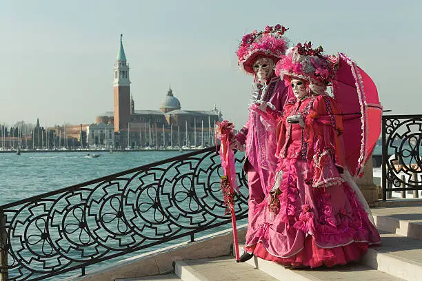 Couple of masks in beautiful purple red costumes on a bridge at carnival in Venice, Italy. Grand Canal and San Giorgio di Maggiore in the background.