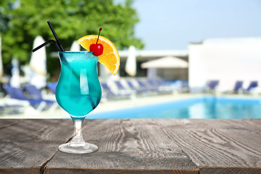 Tasty Blue Lagoon cocktail on wooden table near outdoor swimming pool, space for text