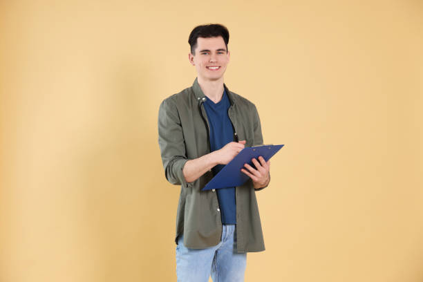 istock Handsome young man writing on clipboard against beige background 1477283372
