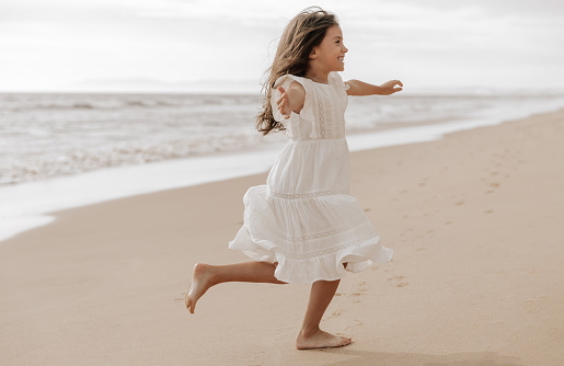 Full body barefoot child in white dress spending arms and smiling while running on sandy beach near waving sea on weekend day