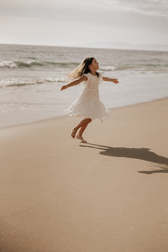 Happy little girl in white dress spreading arms and spinning around while having fun on sandy beach near waving sea in summer