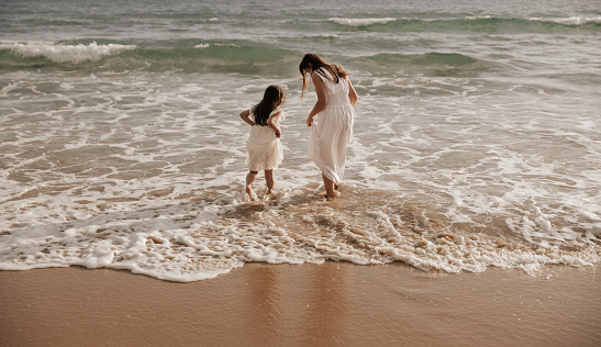 Back view of mother and daughter in white dresses walking into foamy sea waves while spending stormy day on beach together