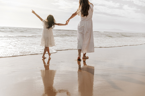 Barefoot kid in white dress pointing at cloudy sky for mom while walking on wet sand near waving sea in morning together