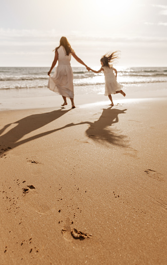 Back view of unrecognizable young woman with long dark hair in maxi white dress holding hand of daughter while running on sandy beach towards wavy ocean at sunset