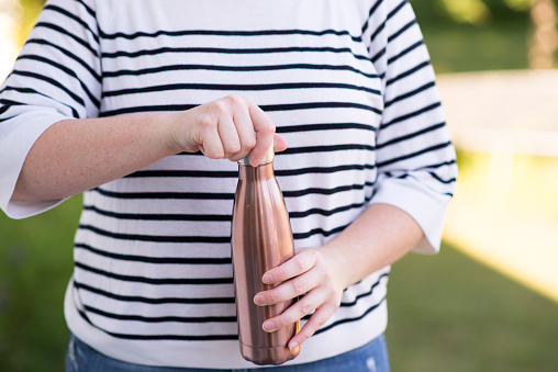 Anonymous young woman is unscrewing the top of a reusable metal water bottle instead of drinking out of disposable plastic water bottles to make it zero waste friendly.
