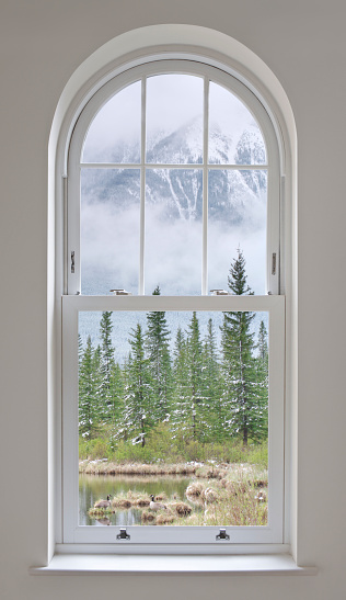 a beautifully made arched window set in a modern new home. The woodwork is in white and the surrounding wall in light grey. The view through the window is over a wintery lake scene, with snow dusted conifers and mountains rising above the mist in the background. Two Canada Geese are in the foreground. This is a composite from two of my images in the iStock Library.