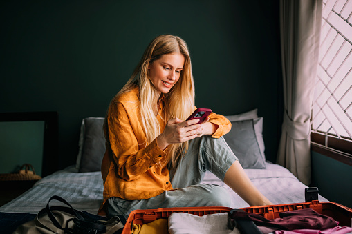 A smiling Caucasian female using her smartphone while sitting on the bed next to her luggage.