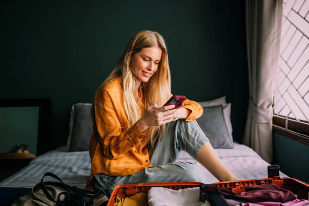 A Happy Beautiful Blonde Businesswoman Texting On Her Mobile Phone While Packing Her Suitcase For A Business Trip stock photo