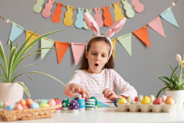 istock Image of amazed excited little girl dressed in rabbit ears sitting at table with Easter eggs, looking with open mouth and surprised face, preparing for celebration, posing against decorated gray wall 1477308223