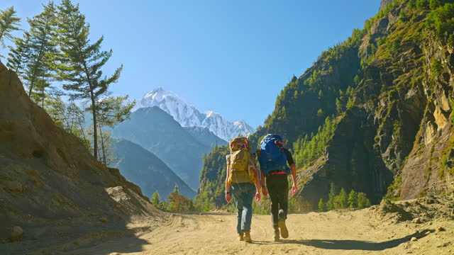 Rear view of trekking pair walking on dusty trail with large and heavy backpacks towards Annapurna mountain range in the Himalayas, Nepal