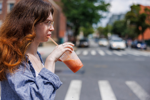 Caucasian young redhead hipster woman in eyeglasses drinking soft juice, while crossing the road in New York City