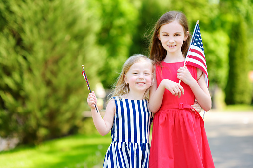 Two adorable little sisters holding American flags outdoors on beautiful summer day. Independence Day concept.