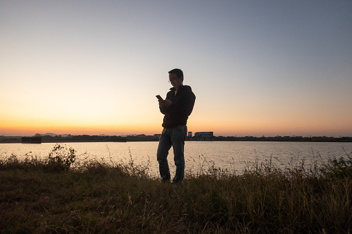 one man alone in the grassland near the river with cell phone after sunset