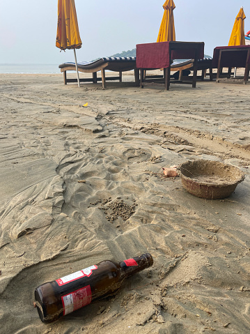 Stock photo showing close-up view of sandy beach at low tide with empty, brown glass beer bottle without lid washed up from polluted sea nearby a row of sun loungers and sunshades in front of water's edge of sea.