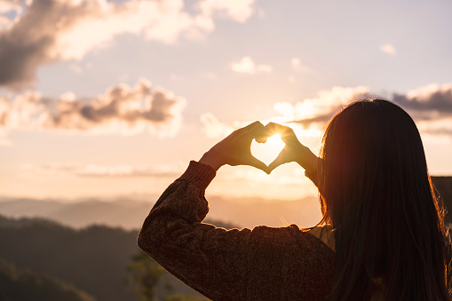 Young woman traveler making heart shape symbol at sunrise over the mountains and looking beautiful landscape, Travel lifestyle concept