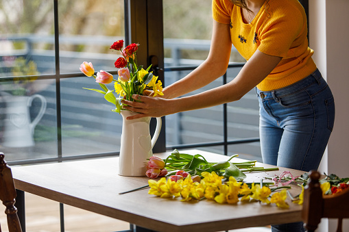 Midsection of unrecognizable, creative young woman putting roses, yellow daffodils and tulips in a vase after arranging it in a bouquet.