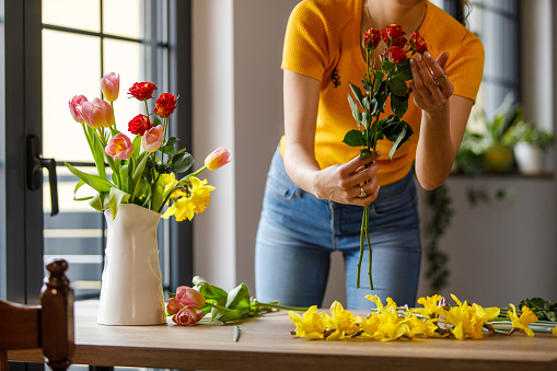 Midsection of unrecognizable young woman standing over a dining table by the window, enjoying arranging yellow daffodils, roses and tulips in a vase.