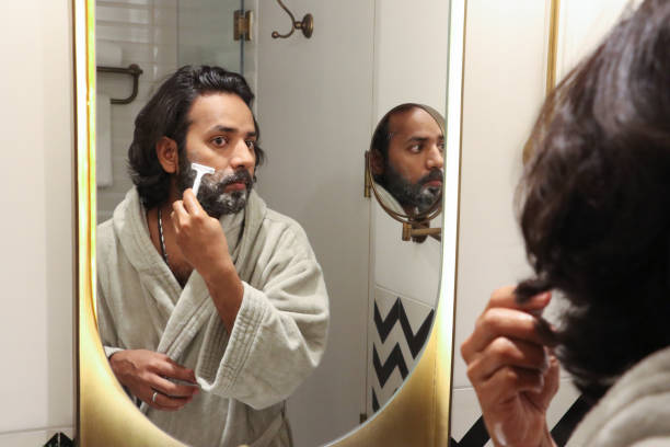 istock Image of reflection in mirror of Indian man stood at sink bathroom wearing bathrobe using razor to trim facial hair, cutting beard and moustache, self care and grooming concept 1477393992