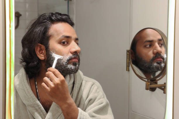 istock Image of reflection in mirror of Indian man stood at sink bathroom wearing bathrobe using razor to trim facial hair, cutting beard and moustache, self care and grooming concept 1477394052
