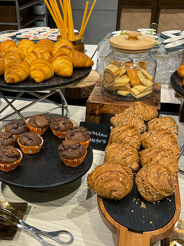 Stock photo showing close-up, elevated view of wood and slate board with rows of freshly baked wholegrain croissants for breakfast ready to be eaten along side a marble cake stand of croissants and a plate of chocolate chip muffins.