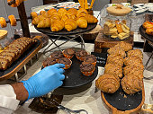 Image of unrecognisable person wearing blue plastic glove to load marble cake stand with chocolate muffins, freshly baked breakfast cakes and pastries at hotel restaurant buffet, row of wholegrain croissants on wood and slate board, glass cookie jar