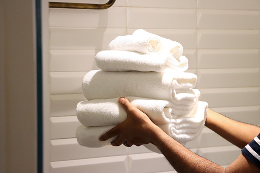 Stock photo showing close-up view of pile of folded, white towels being carried by an unrecognisable person to put on high shelf in a luxury hotel bathroom with horizontal stacked pattern, white bevelled edged wall tiles. Home furnishings and bedroom interior design.