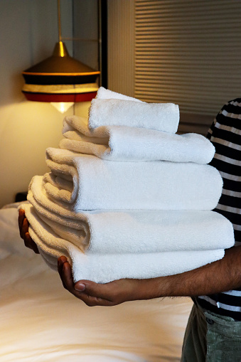 Stock photo showing close-up view of pile of folded, white towels being carried by an unrecognisable person in a hotel room with a double bed covered with white duvet bedding with background of gold cushions and a stack of white hotel pillows resting on textured, baton headboard. Home furnishings and bedroom interior design.

Close-up image of unrecognisable person carrying stack of white hotel towels, folded bath sheets and hand towels, white duvet double bed cover, circular, yellow velour cushion leaning on pile of white pillows against textured, baton headboard