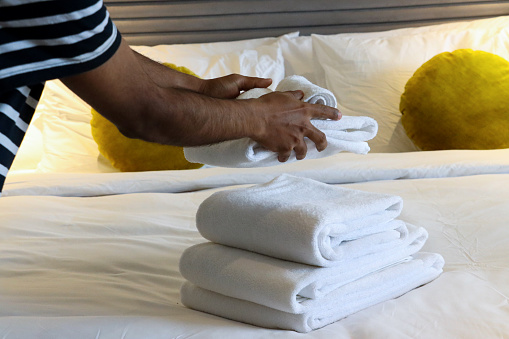 Stock photo showing close-up view of pile of folded, white towels sitting on hotel room double bed duvet bedding with background of gold cushions and a stack of white hotel pillows resting on textured, baton headboard. Home furnishings and bedroom interior design.